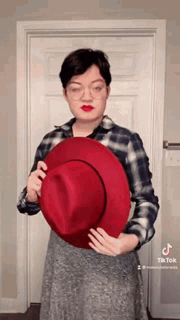 a woman wearing a plaid shirt and red lipstick is holding a red hat .