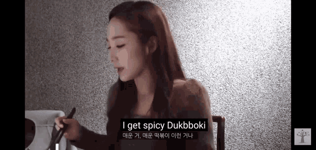 a woman is sitting in a chair holding a knife and saying `` i get spicy dukbboki ''
