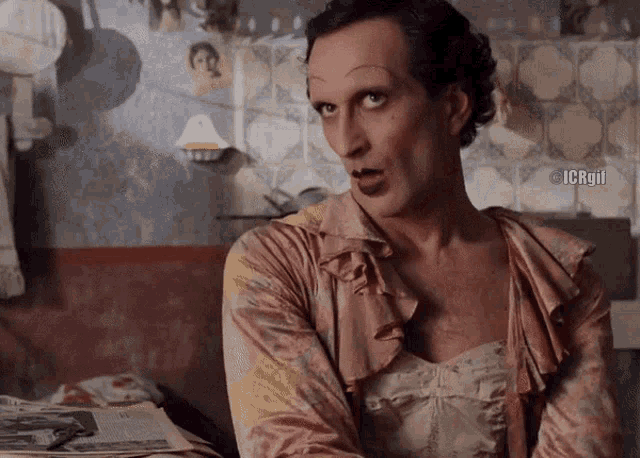 a man dressed as a woman is sitting in front of a wall with pictures on it
