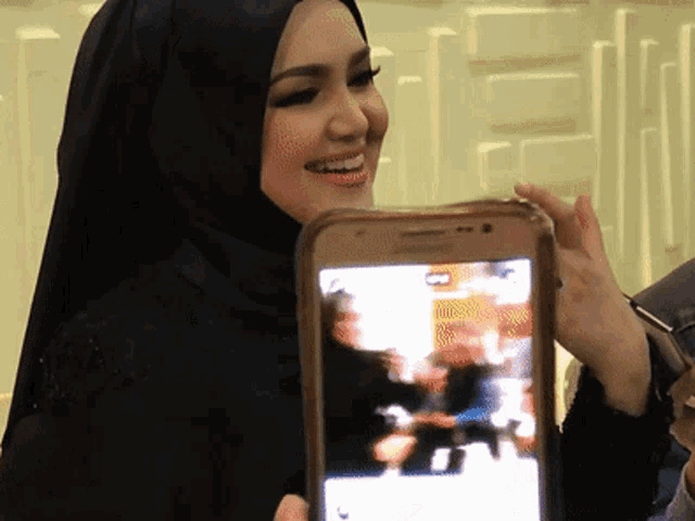 a woman in a black hijab smiles while holding a phone