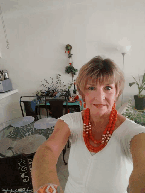 a woman wearing a white shirt and a red necklace takes a selfie in front of a table
