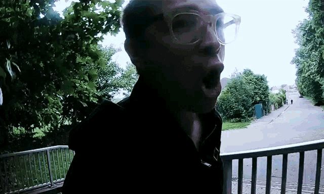 a man wearing glasses stands in front of a fence with his mouth open