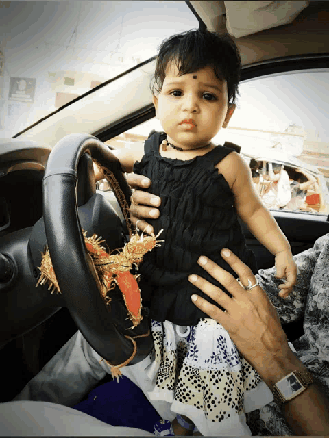 a little girl in a black dress is sitting in a car with a steering wheel cover on it