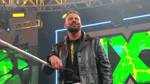 a man in a leather jacket is standing in a wrestling ring with a green x in the background
