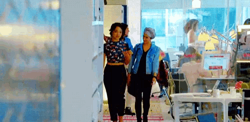 two women are walking down a hallway in an office with a man sitting at a desk .