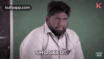 a man with a beard is sitting in front of a chalkboard and saying `` shocked '' .