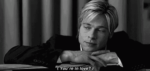 a man in a suit is sitting at a table with his head resting on his hands and says " you 're in love "