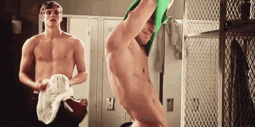 two shirtless men are standing in a locker room and one of them is taking off his shirt .