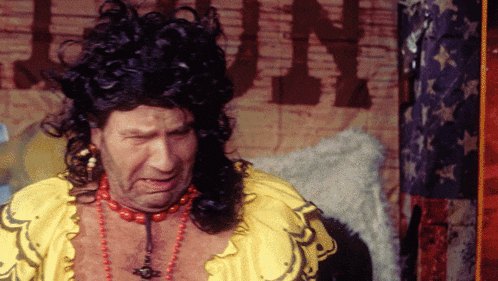 a man wearing a wig and a necklace stands in front of a sign that says ' guns ' on it