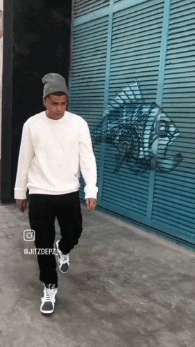 a man wearing a beanie and a white sweater is walking in front of a blue wall