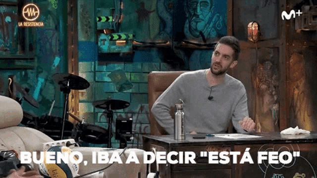 a man is sitting at a desk in front of a drum set and says bueno iba a decir esta feo .
