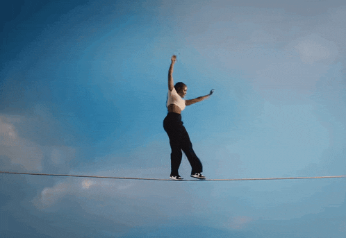a woman is walking on a tightrope with a blue sky behind her