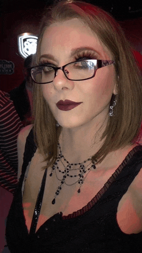 a woman wearing glasses and a necklace looks at the camera with a red background