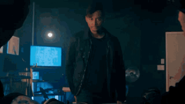 a man in a black jacket is standing in a dark room with a computer monitor in the background .