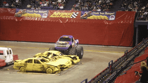 a monster truck is driving through a pile of wrecked cars in front of an advance auto parts sign
