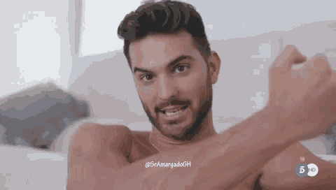 a shirtless man with a beard is laying on a bed with his arms crossed and smiling .