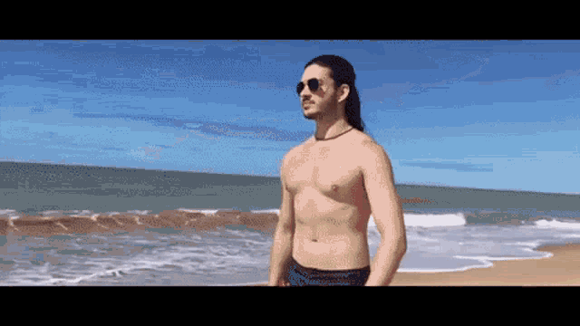 a shirtless man with long hair stands on a beach