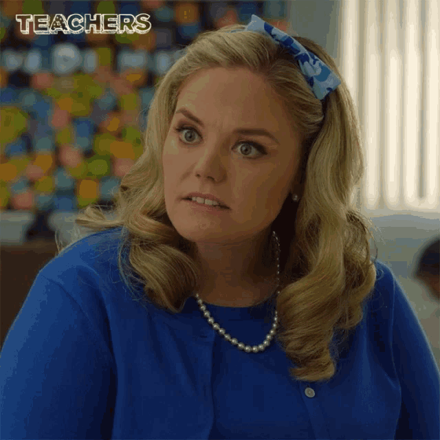 a woman wearing a blue shirt and a pearl necklace is sitting in front of a bulletin board that says teachers .