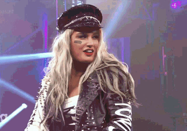 a blonde woman wearing a black hat and a leather jacket stands in front of a stage