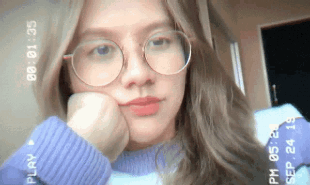 a woman wearing glasses and a purple sweater has a play button on her sweater