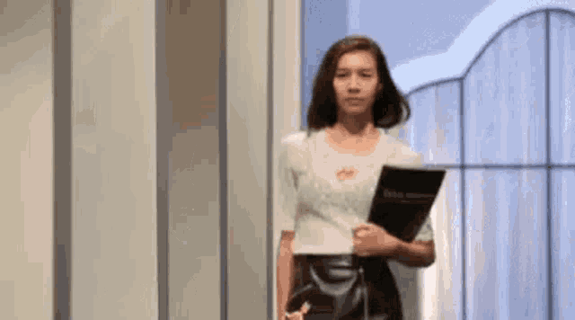 a woman in a white top and black skirt is standing in a hallway holding a folder .