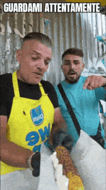 two men are standing next to each other eating corn on the cob . one of the men is wearing a yellow apron .
