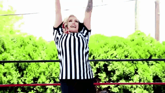 a woman in a striped referee shirt is standing in a ring with her arms in the air .