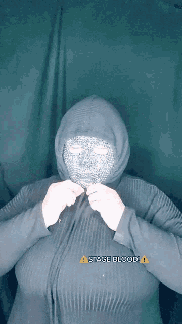 a woman wearing a mask and a hoodie has stage blood written on the bottom