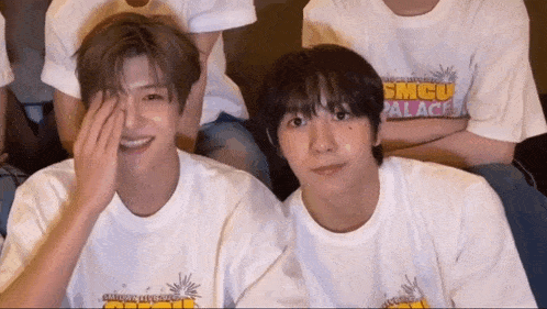 two young men wearing white shirts with smcu palace on them are sitting next to each other .