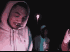 a man in a hoodie is smoking a cigarette in the dark while another man looks on .