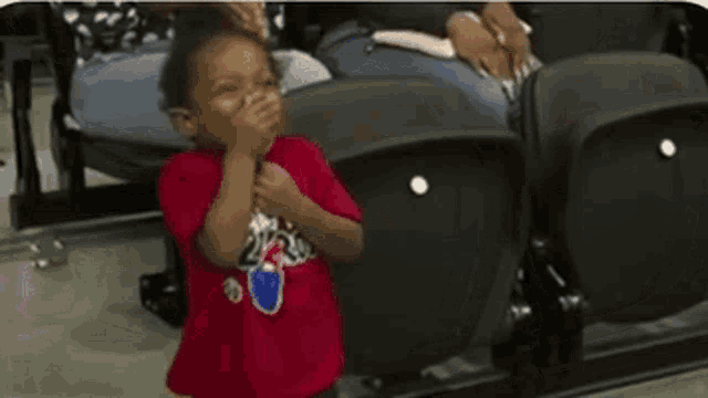 a little boy in a red shirt is sitting in a stadium covering his mouth with his hand .