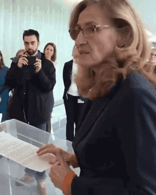 a woman wearing glasses holds a piece of paper in front of a group of people
