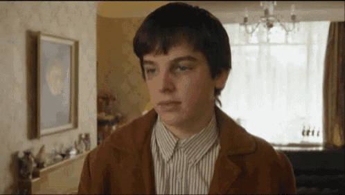 a young boy wearing a brown jacket and striped shirt is standing in a living room