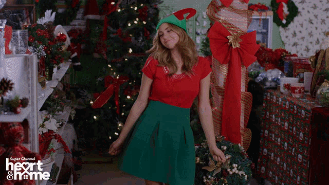a woman in a red top and green skirt is dancing in front of a christmas tree with a super channel logo on the bottom