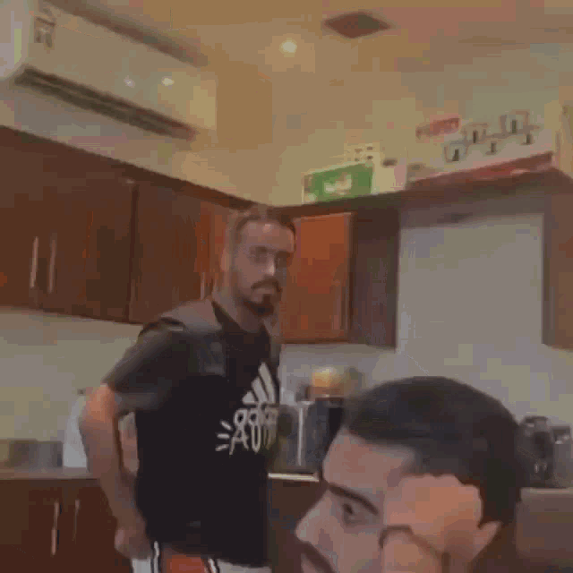 a man with a beard and glasses is standing in a kitchen with another man .