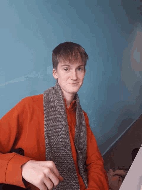 a young man wearing an orange sweater and a scarf looks at the camera