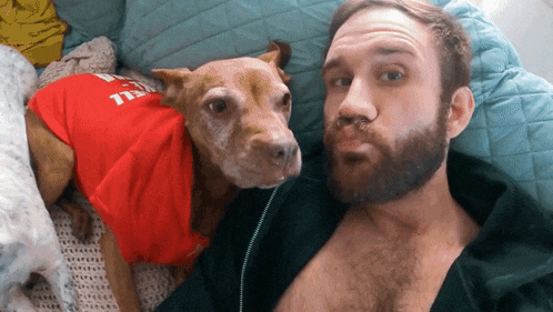 a man with a beard is laying next to a dog wearing a red shirt that says ' hell ' on it