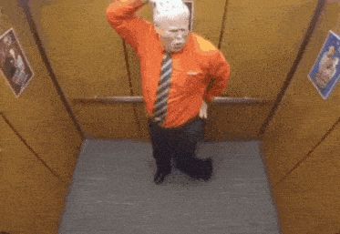 a man in an orange shirt and tie is standing in an elevator giving a thumbs up .