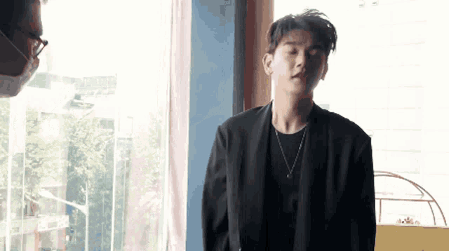 a man wearing a black jacket and a necklace is standing in front of a window