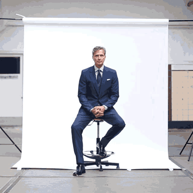 a man in a blue suit sits on a stool in front of a white background