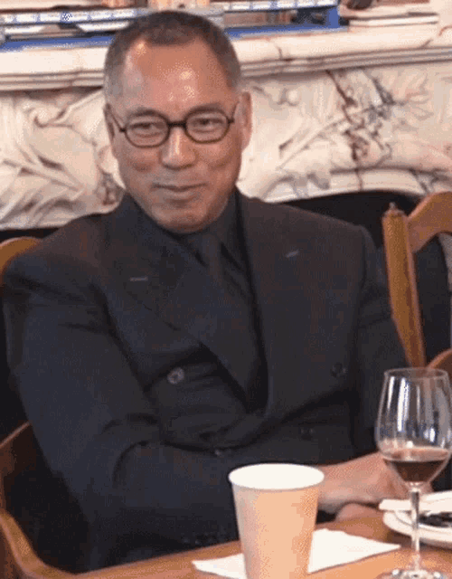 a man in a suit sits at a table with a glass of wine