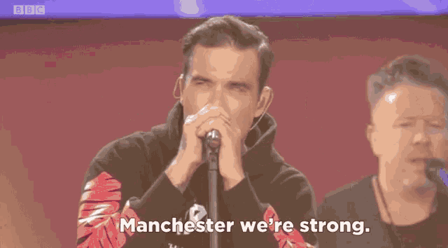 a man singing into a microphone with the words manchester we 're strong behind him