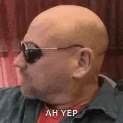 a bald man wearing sunglasses and a green shirt is sitting down .