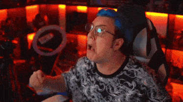 a man with blue hair and glasses is sitting in a chair in front of a computer screen .