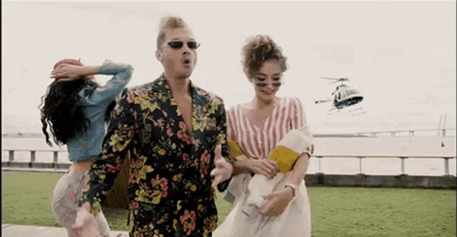 a man in a floral suit is standing next to two women while a helicopter flies overhead .