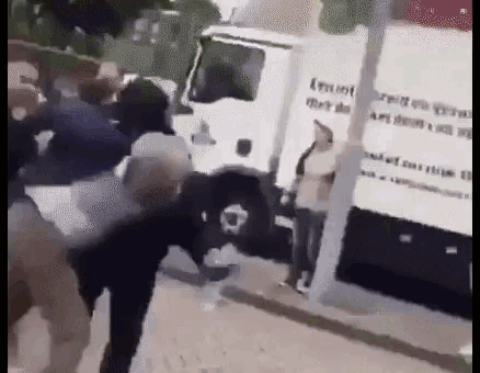 a group of people are fighting in front of a white truck on a street .