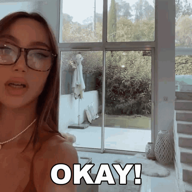a woman with glasses says okay in front of a window