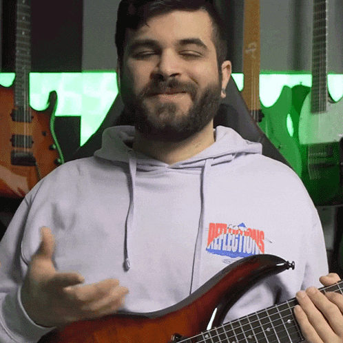 a man wearing a reflections hoodie holds a guitar