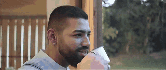 a man with a beard is drinking from a cup while looking out of a window .