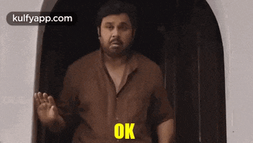 a man in a brown shirt is standing in front of a door and saying `` ok '' .